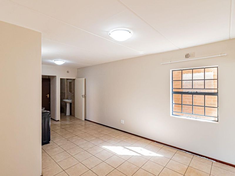 An empty, neutrally painted living area featuring tiled flooring, flush mount lighting, and a window with security bars, leading to an adjacent bathroom space.