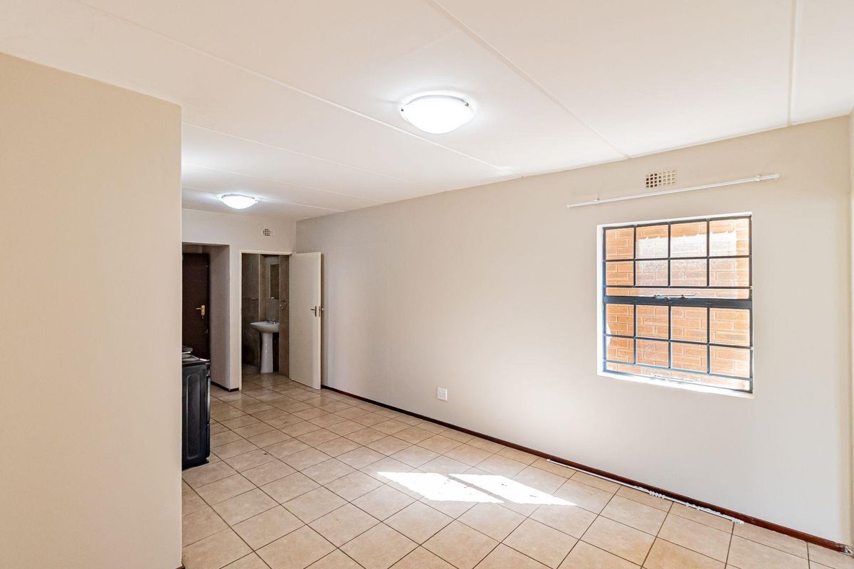 An empty, neutrally painted living area featuring tiled flooring, flush mount lighting, and a window with security bars, leading to an adjacent bathroom space.