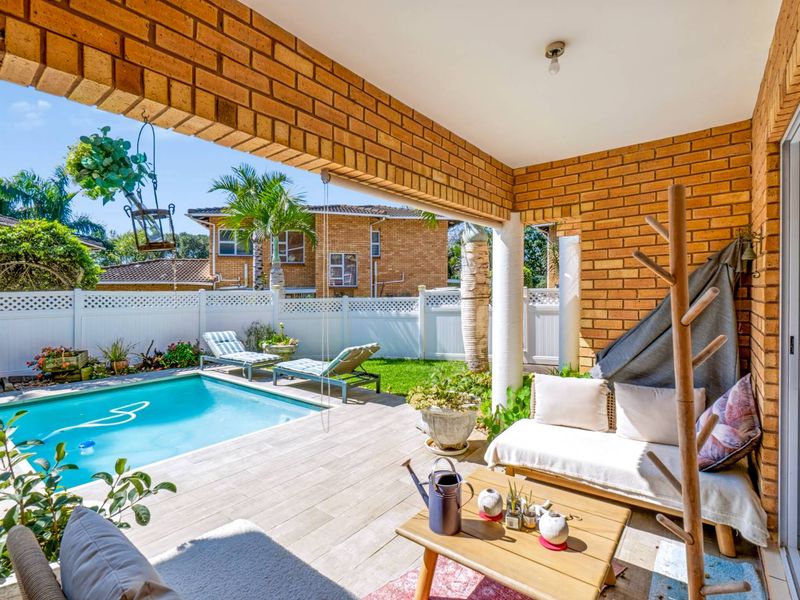 Covered patio area featuring exposed brickwork and tiled flooring, overlooking a private swimming pool and garden with white perimeter fencing.