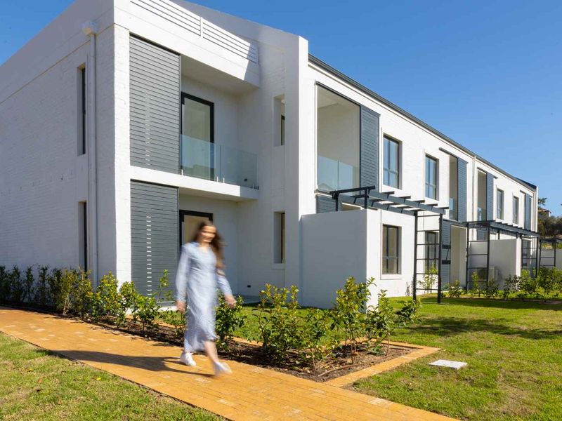 Modern apartment exterior featuring clean white facades, glass balcony balustrades, and a well-maintained landscaped garden with a paved walkway.