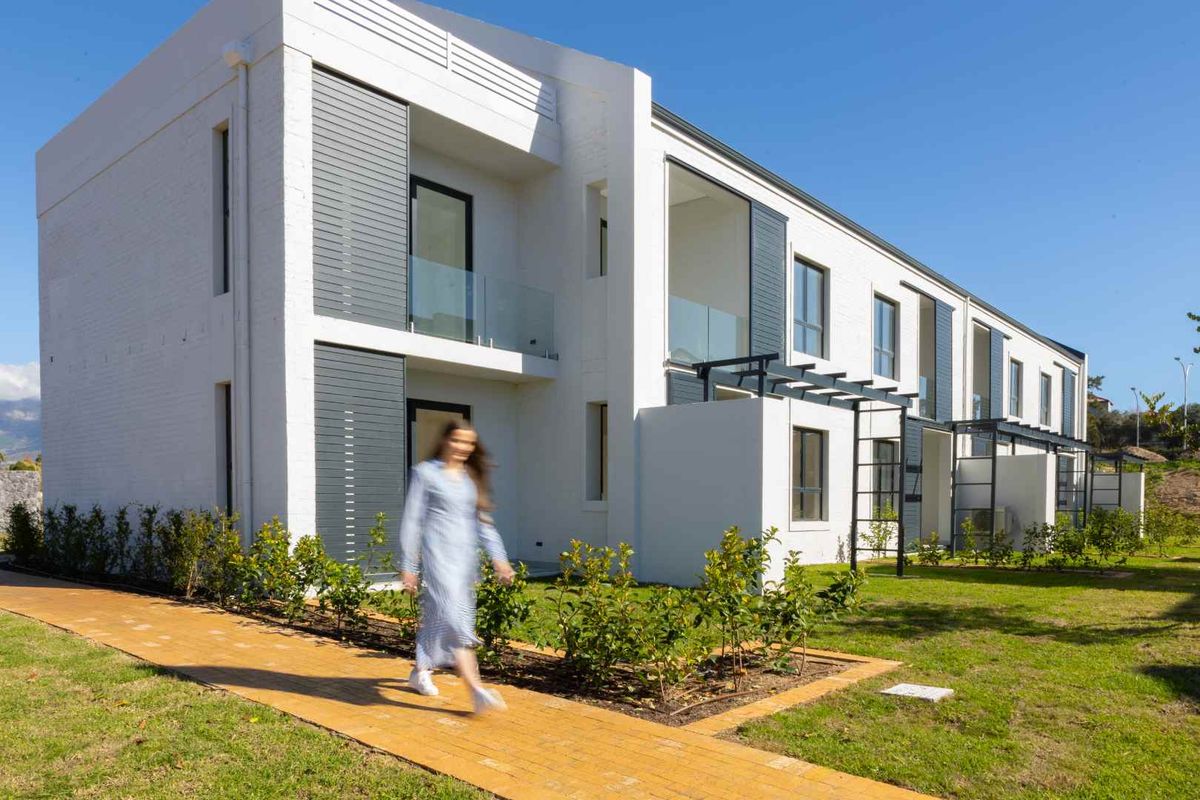 Modern apartment exterior featuring clean white facades, glass balcony balustrades, and a well-maintained landscaped garden with a paved walkway.