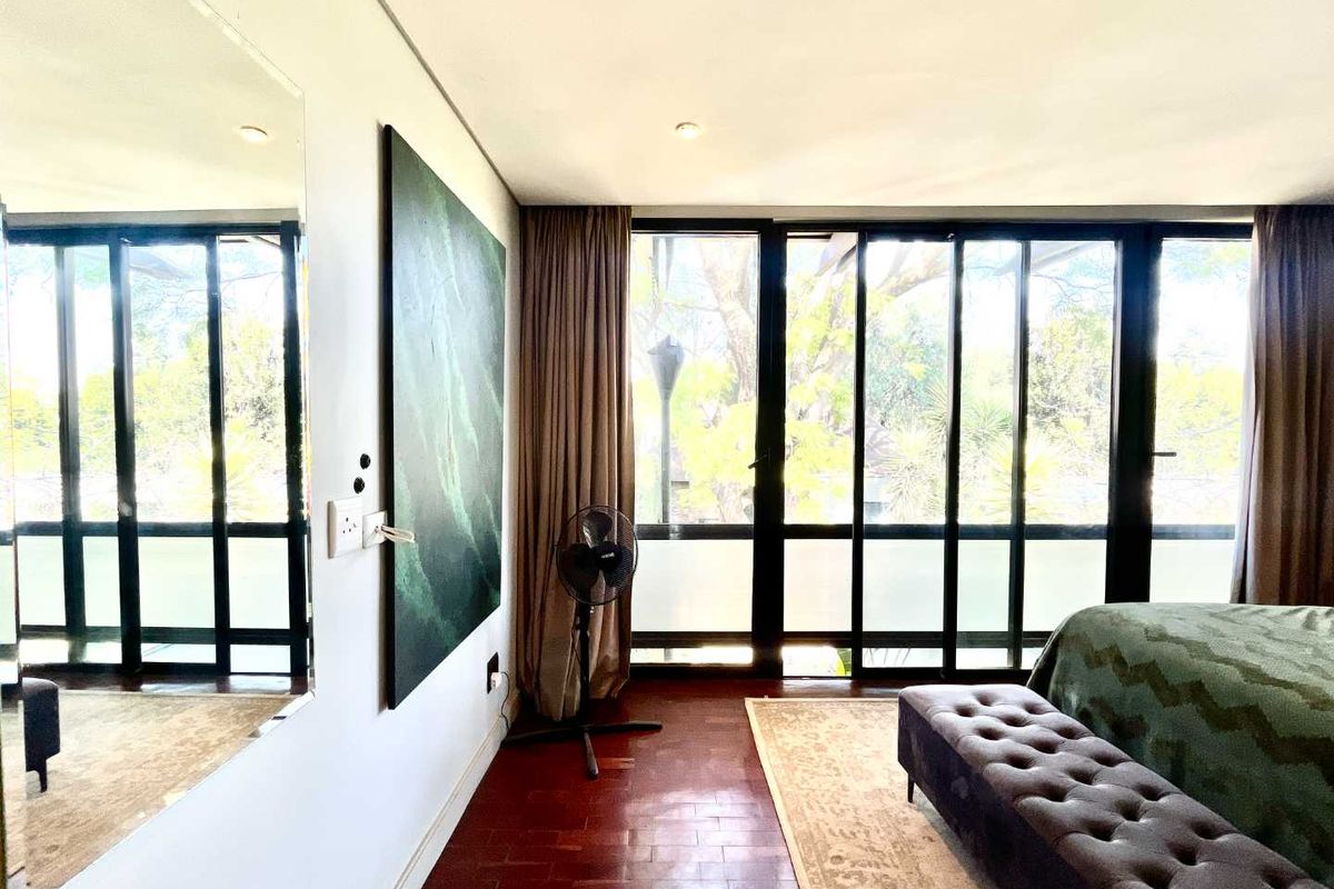 A bright bedroom featuring warm parquet flooring, large floor-to-ceiling windows that provide ample natural light, and a wall-mounted mirror.
