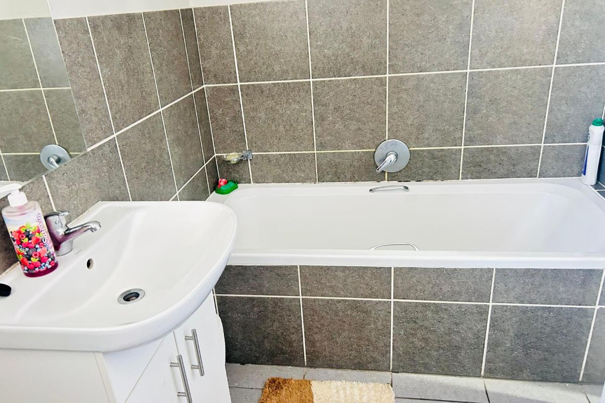 A functional bathroom featuring a white vanity basin, a built-in bathtub with chrome fixtures, and grey tiled walls.