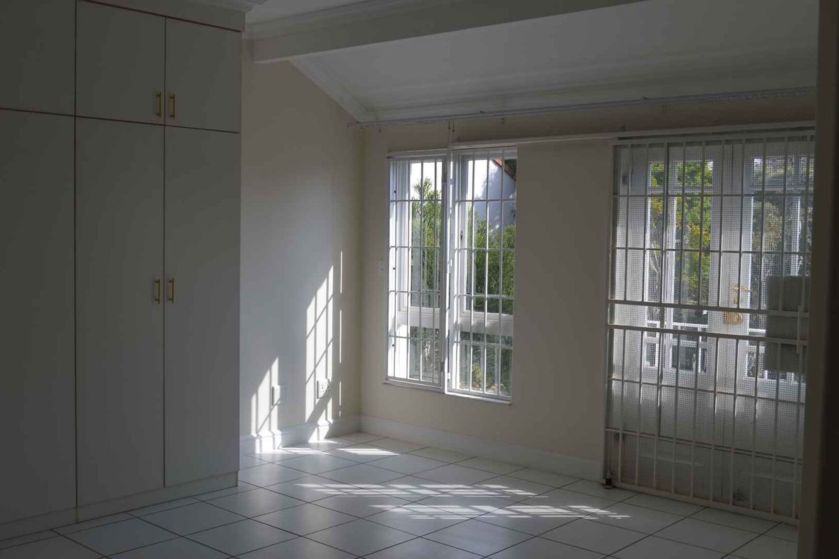 A bright, empty bedroom featuring tiled flooring, built-in cupboards, and secure window and door access with fitted security bars and gates.