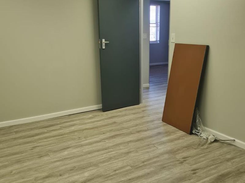 A clean, vacant office space featuring modern light-wood laminate flooring, neutral walls, and a suspended ceiling with recessed lighting.