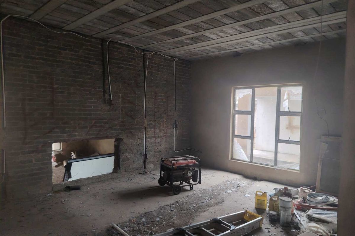 An unfinished living room space featuring exposed brickwork, concrete ceiling beams, and an aluminium window frame, currently undergoing renovation.