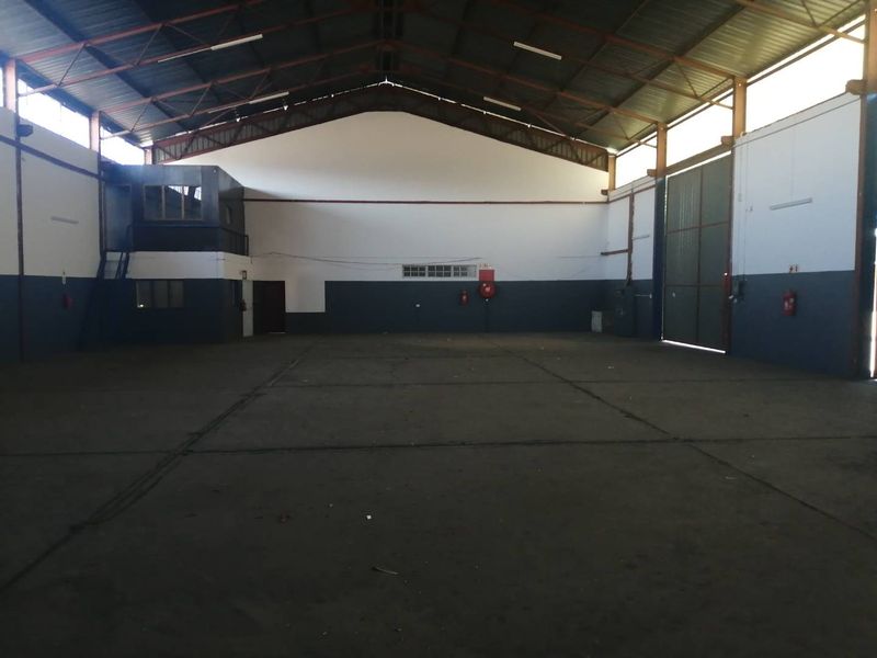Spacious industrial warehouse featuring high ceilings with exposed steel trusses, concrete flooring, a mezzanine office area, and a large sliding access door.