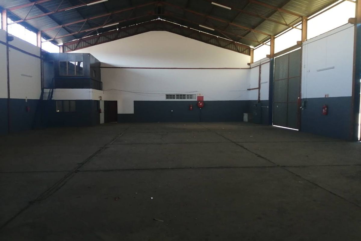 Spacious industrial warehouse featuring high ceilings with exposed steel trusses, concrete flooring, a mezzanine office area, and a large sliding access door.