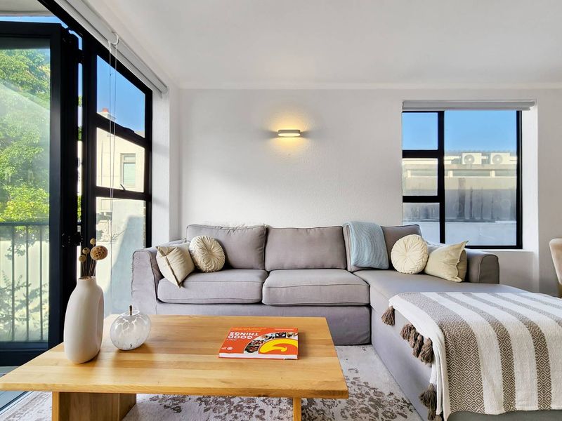 A bright and modern living room featuring large black-framed windows, a sliding door to the balcony, and a minimalist wall-mounted light fixture.