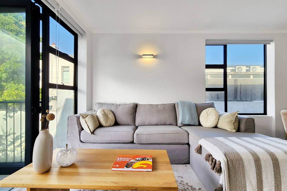 A bright and modern living room featuring large black-framed windows, a sliding door to the balcony, and a minimalist wall-mounted light fixture.