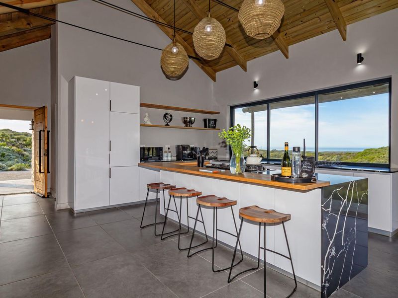 Modern open-plan kitchen featuring a large island with stone countertop, exposed timber rafters, and expansive sliding doors offering scenic coastal views.