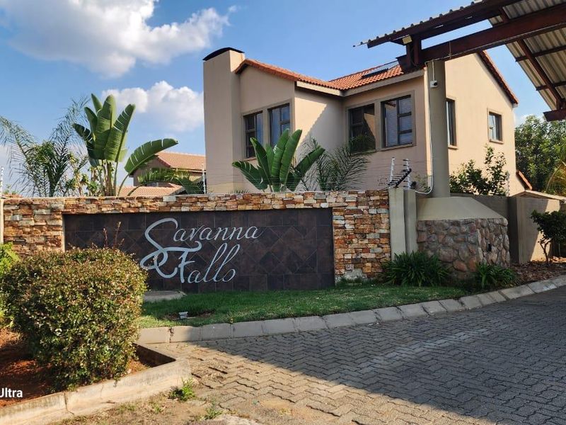 Front entrance view of the Savanna Falls townhouse complex featuring stone-clad boundary walls, paved driveway, and secure gated access.