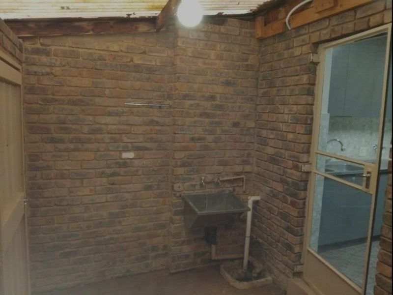A functional laundry area featuring exposed brick walls, a stainless steel utility sink, and basic plumbing connections under a corrugated roof structure.