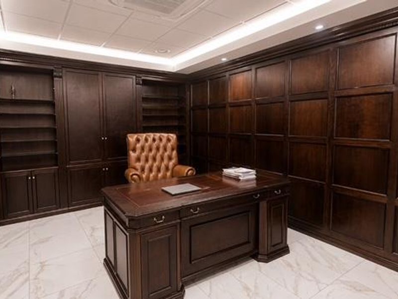 A sophisticated home office featuring extensive dark wood built-in cabinetry, elegant wood wall paneling, and polished marble-look floor tiles with recessed ceiling lighting.