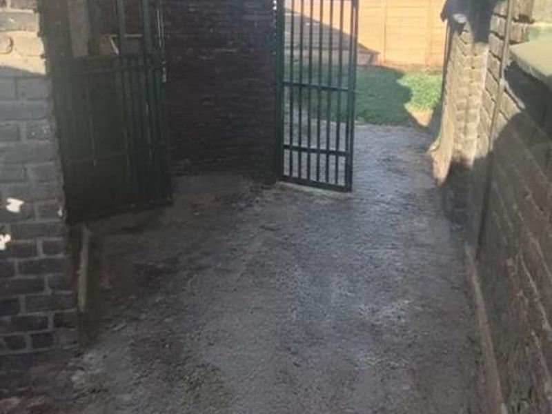 A narrow rear exterior passage featuring brick walls, a corrugated metal roof covering, and a metal security gate leading to the backyard.