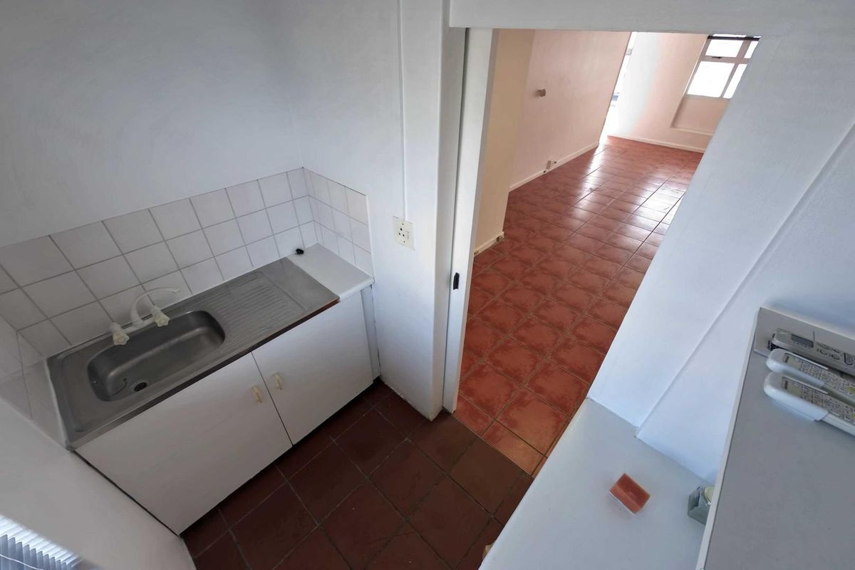 A compact kitchenette area featuring a stainless steel sink, tiled splashback, and built-in cabinetry, leading into an adjacent tiled office space.