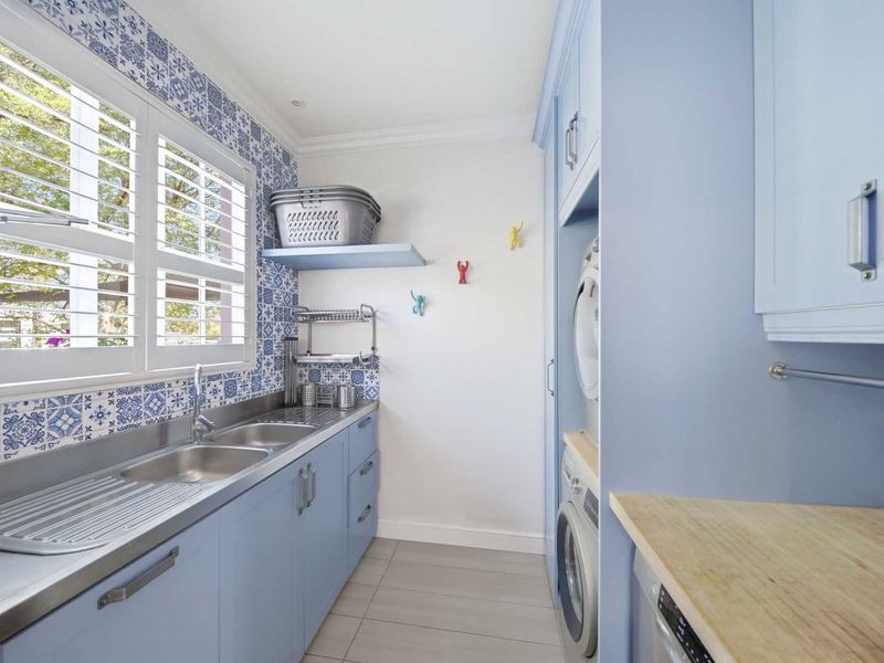 A well-appointed laundry room featuring blue cabinetry, a stainless steel double sink, patterned tiled splashback, and plantation shutters.