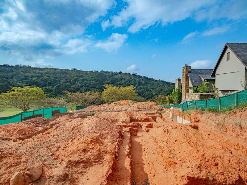 A vacant residential plot showing excavated foundation trenches, ready for construction, with adjacent established housing and scenic hillside views.