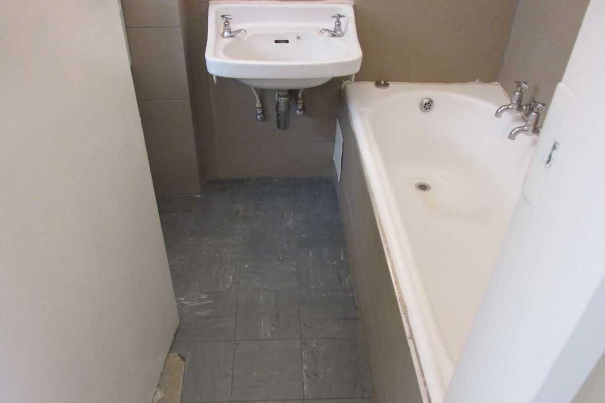 A compact bathroom featuring a wall-mounted basin and a built-in bathtub with tiled walls and vinyl flooring, currently in need of cosmetic updates.