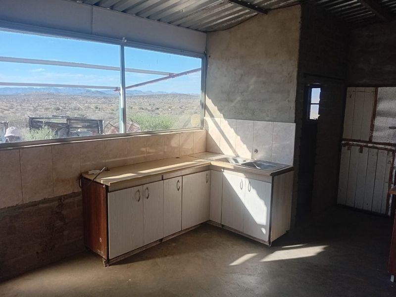 A rustic kitchen area featuring built-in cabinetry, a double sink, tiled splashbacks, and expansive views through a large window, set within an unfinished interior space.