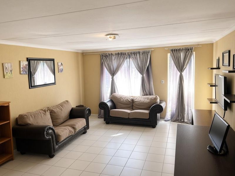 A spacious living room featuring neutral tiled flooring, soft yellow walls, and natural light through large windows with curtain fixtures.
