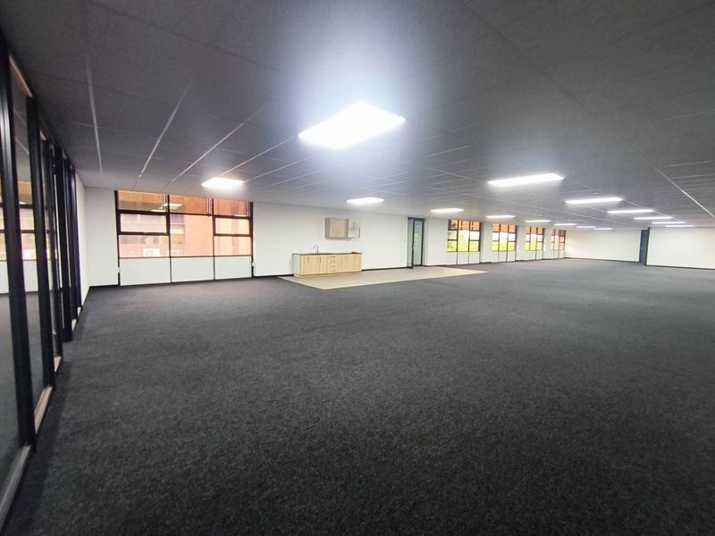 A spacious, open-plan office area featuring dark carpeting, a suspended ceiling with recessed lighting, and a small built-in kitchenette unit.