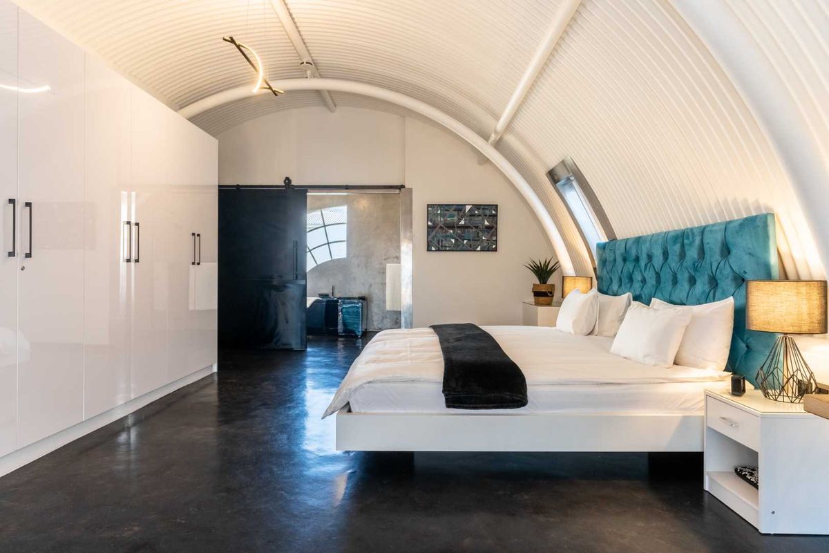 Modern bedroom featuring a unique curved corrugated metal ceiling, polished concrete floors, and extensive built-in white cabinetry with a sleek sliding door entry.