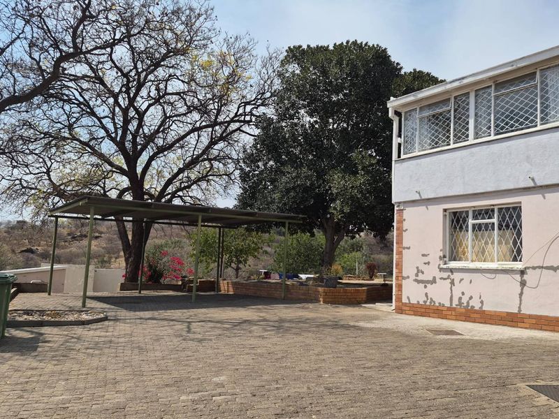 Rear exterior view of the property featuring a paved driveway, a metal carport, and mature trees, showing signs of exterior wall maintenance required.