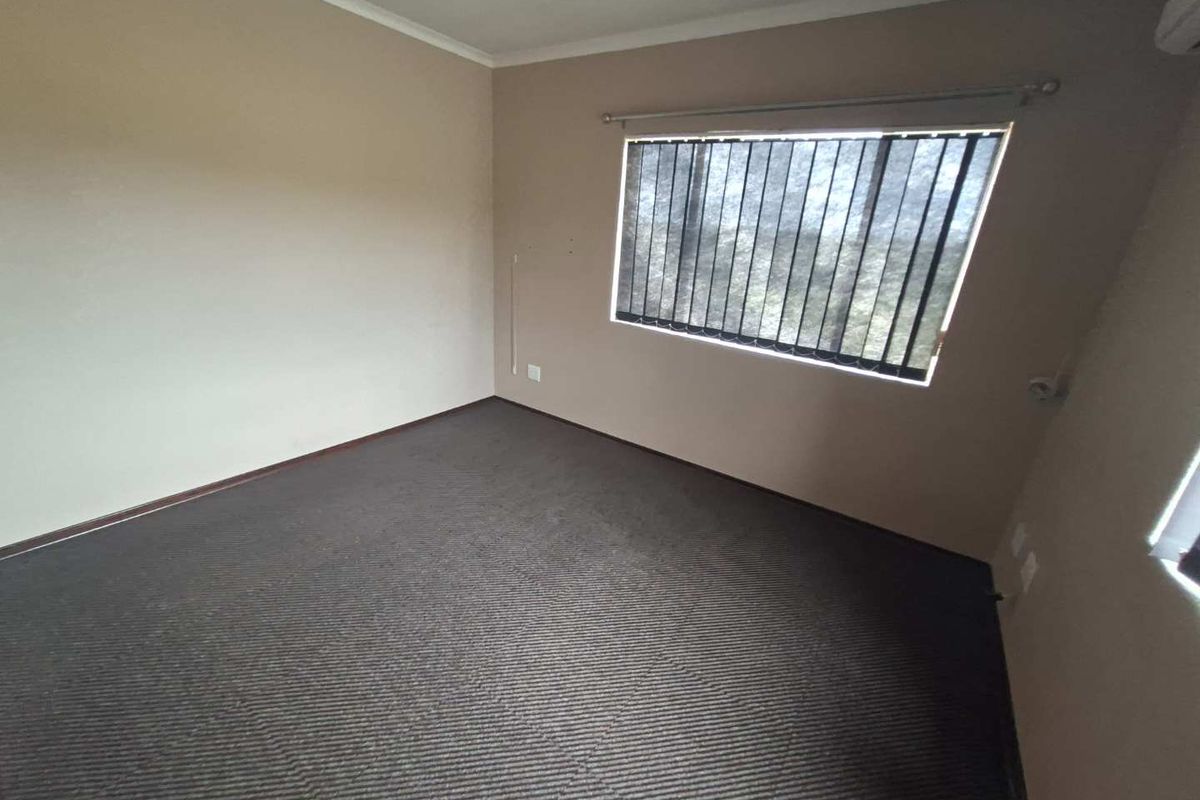 An empty bedroom featuring neutral wall paint, dark carpeting, vertical window blinds, and a wall-mounted air conditioning unit.