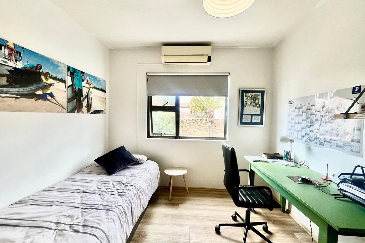 A functional bedroom featuring laminate flooring, a wall-mounted split-system air conditioner, and a window with a roller blind.