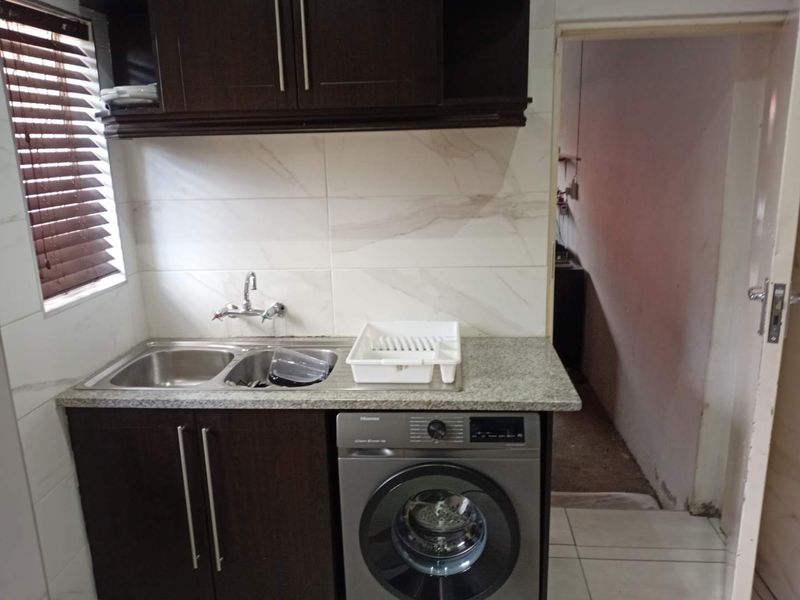 A functional laundry area featuring dark wood cabinetry, granite countertops, a double sink, and tiled backsplash, with an adjacent utility space.