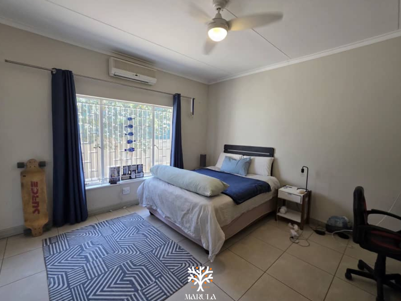 A functional bedroom featuring tiled flooring, a ceiling fan, and a wall-mounted air conditioning unit, with security bars installed on the window.