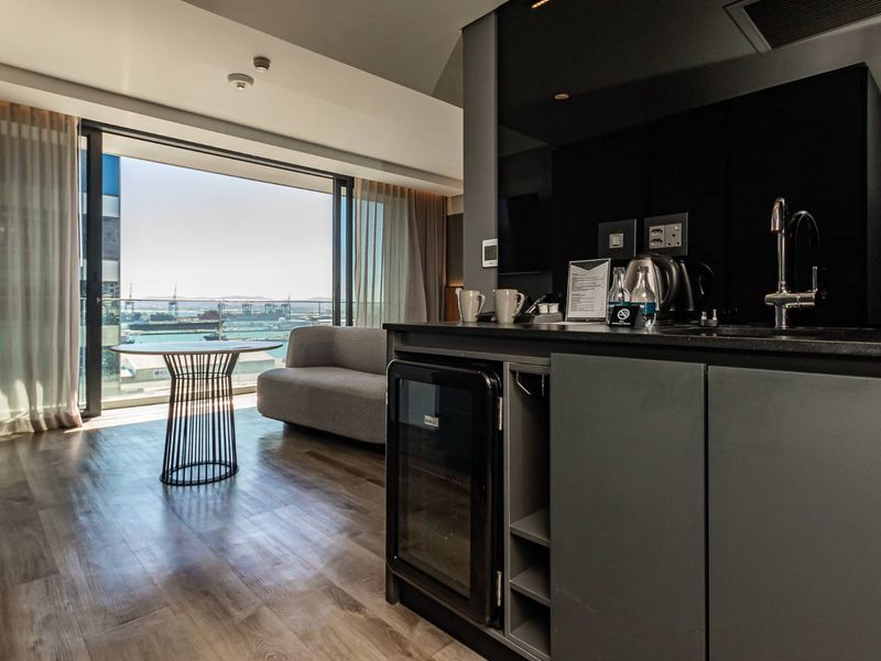 Modern open-plan apartment featuring a sleek kitchenette with integrated cabinetry, wood-look flooring, and floor-to-ceiling glass doors opening to a balcony with harbor views.