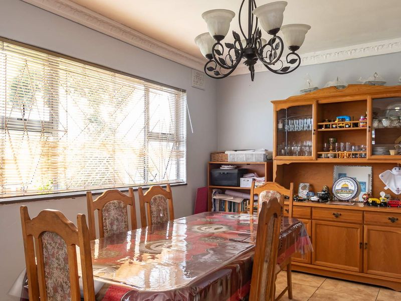 A dining room featuring tiled flooring, an ornate ceiling cornice, and a large window with venetian blinds, illuminated by a decorative wrought iron chandelier.