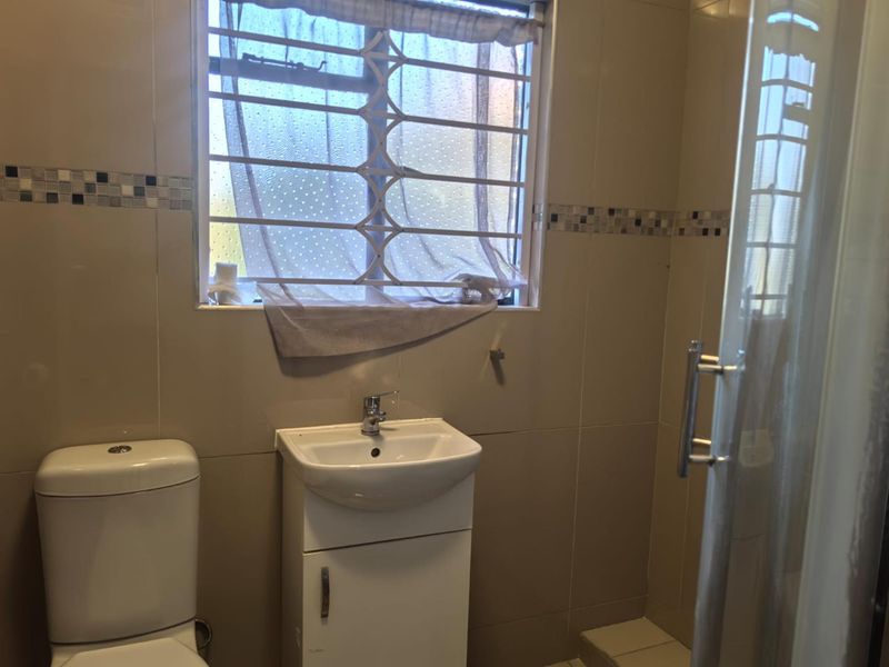 A functional bathroom featuring a vanity cabinet, ceramic basin, toilet, and a shower enclosure with tiled walls and security bars on the window.