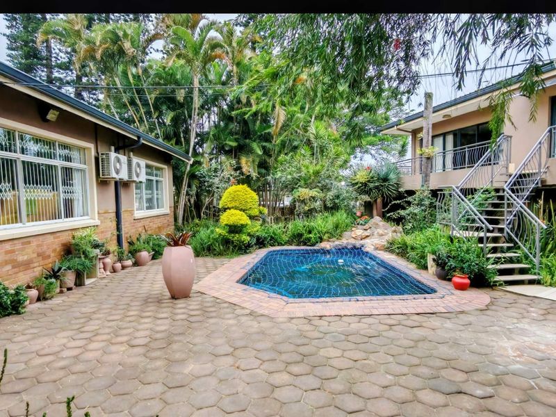 A well-maintained outdoor pool area featuring a paved patio, established garden landscaping, and an external staircase leading to an upper level.