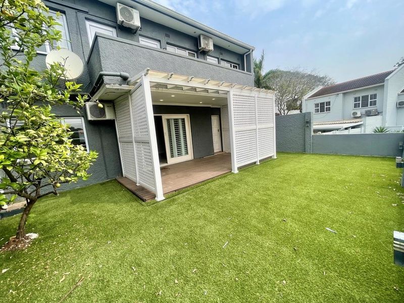 Rear view of the townhouse featuring a covered patio with timber decking, privacy trellis, and a well-maintained artificial lawn garden area.