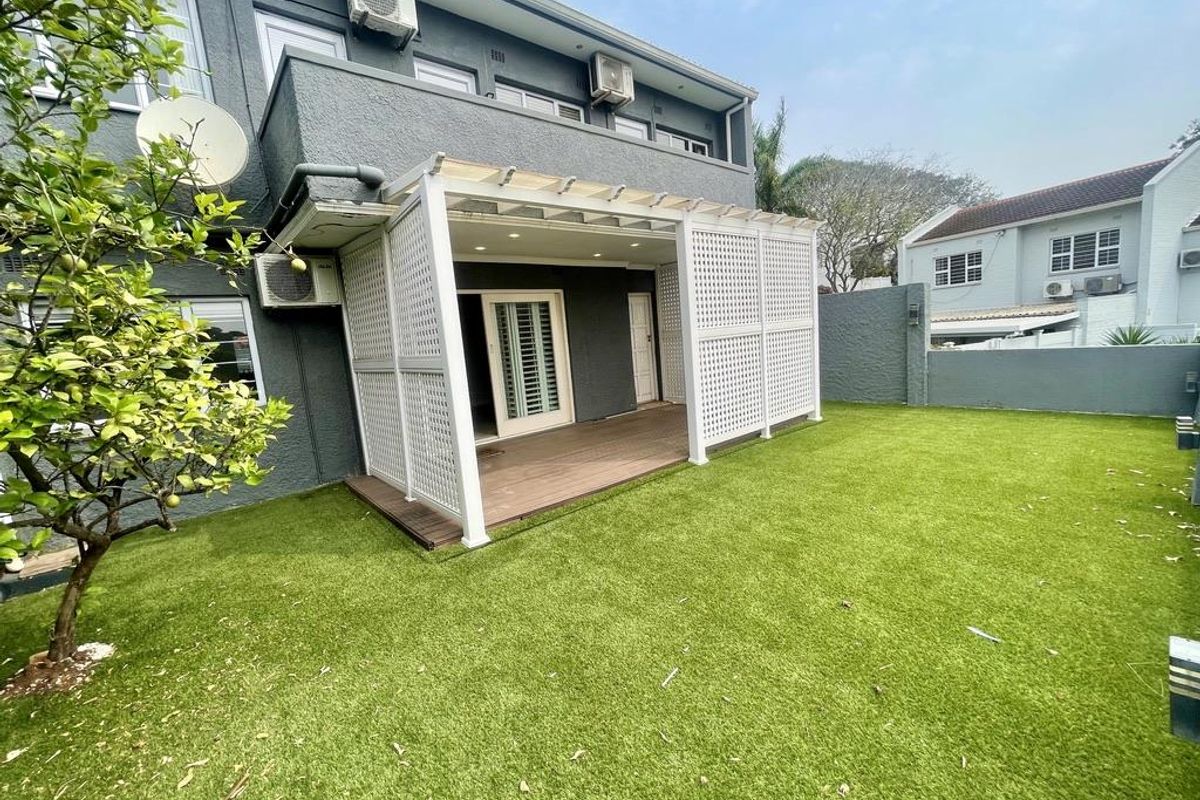 Rear view of the townhouse featuring a covered patio with timber decking, privacy trellis, and a well-maintained artificial lawn garden area.