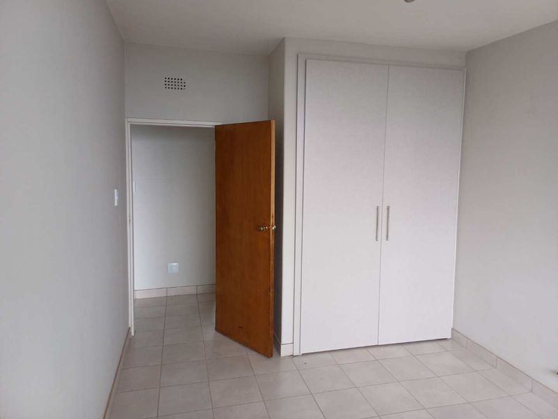 A clean, empty bedroom featuring neutral tiled flooring, a built-in white wardrobe, and a wooden door leading to an adjacent space.