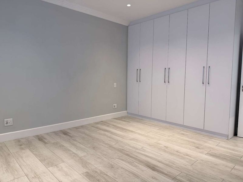 A bright, empty bedroom featuring modern light-toned laminate flooring, neutral grey walls, and extensive floor-to-ceiling built-in cupboards.