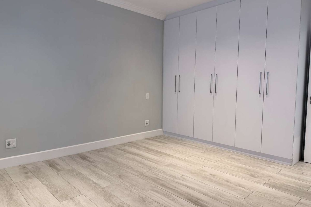 A bright, empty bedroom featuring modern light-toned laminate flooring, neutral grey walls, and extensive floor-to-ceiling built-in cupboards.
