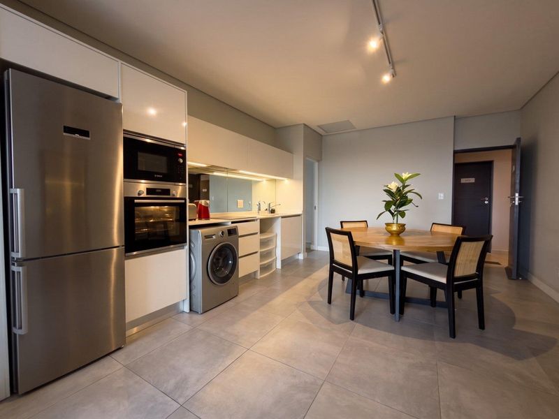 Modern open-plan kitchen and dining area featuring sleek white cabinetry, integrated appliances, mirrored splashback, and neutral tiled flooring throughout.