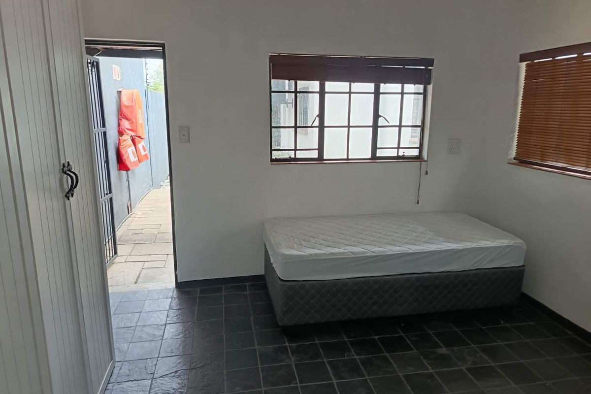 A simple bedroom featuring dark slate-style floor tiles, white walls, and a window with wooden blinds, providing direct access to an outdoor area.