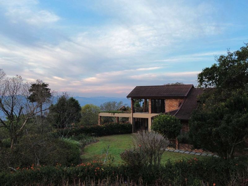 An elevated rear view of a brick-faced house featuring a covered patio, balcony, and expansive landscaped garden with scenic mountain views.