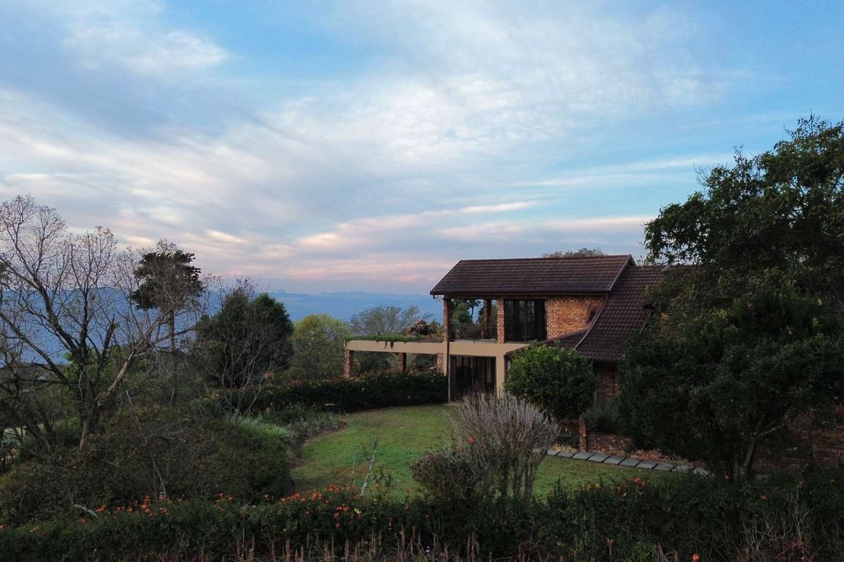 An elevated rear view of a brick-faced house featuring a covered patio, balcony, and expansive landscaped garden with scenic mountain views.