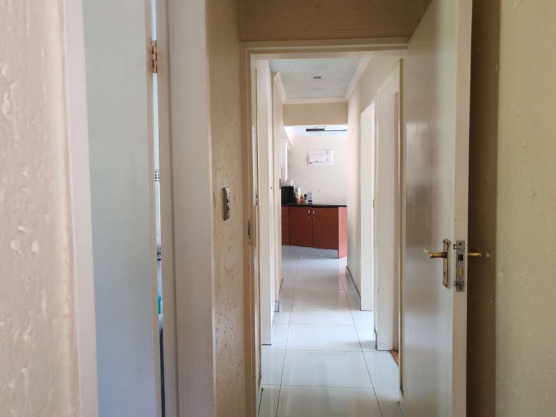 A view through a residential hallway with tiled flooring, leading towards a kitchen area, showing signs of wear on walls and door frames.