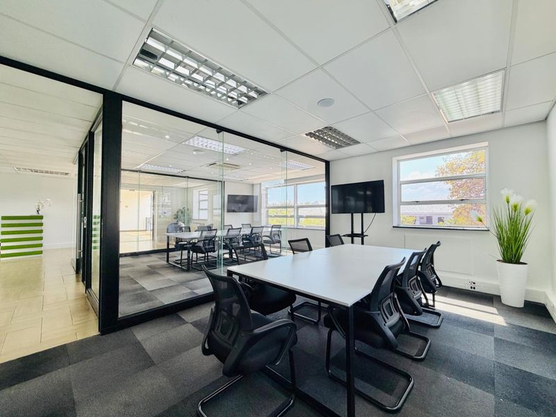 Modern office space featuring a glass-partitioned meeting room, carpeted flooring, suspended ceilings with recessed lighting, and large windows providing natural light.