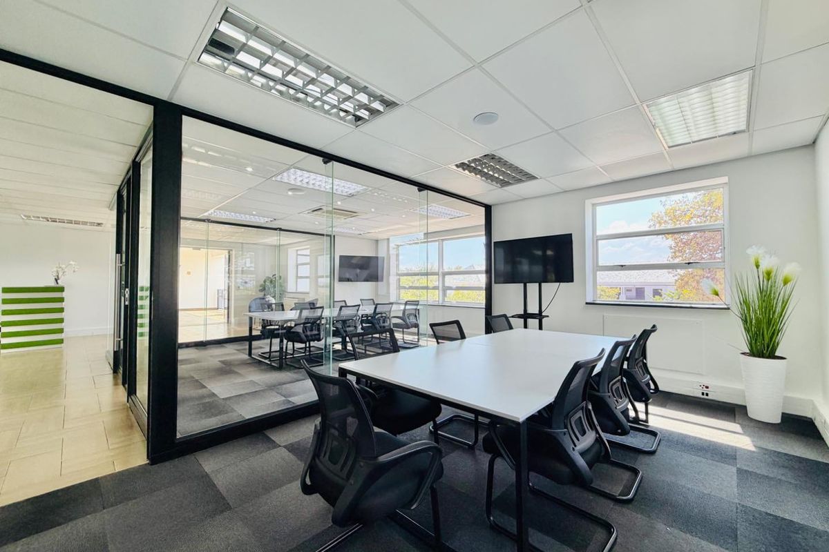 Modern office space featuring a glass-partitioned meeting room, carpeted flooring, suspended ceilings with recessed lighting, and large windows providing natural light.