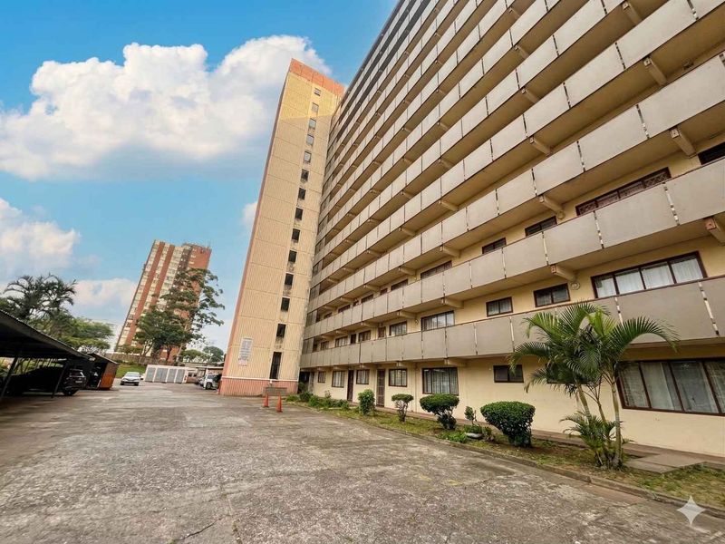 Exterior view of a multi-story apartment building featuring concrete balconies and an adjacent paved parking area.