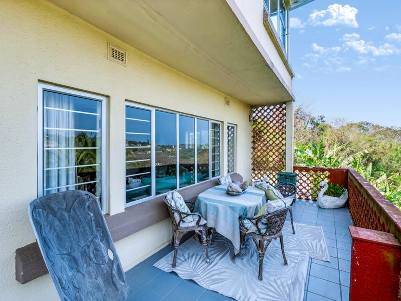 A covered balcony featuring tiled flooring, security bars on windows, and a brick balustrade, offering an outdoor space with views of the surrounding greenery.